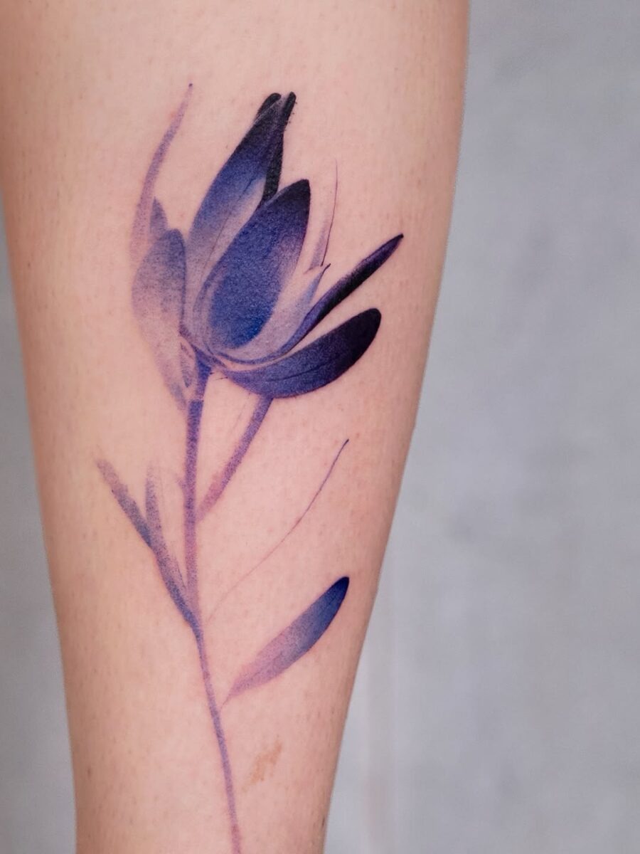 This tattoo features a striking watercolor design of a flower, blending deep purples and soft pinks for a dreamy, ethereal vibe.