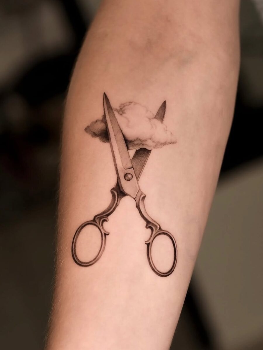 This unique tattoo features a pair of intricately designed scissors crossing through a delicate cloud.