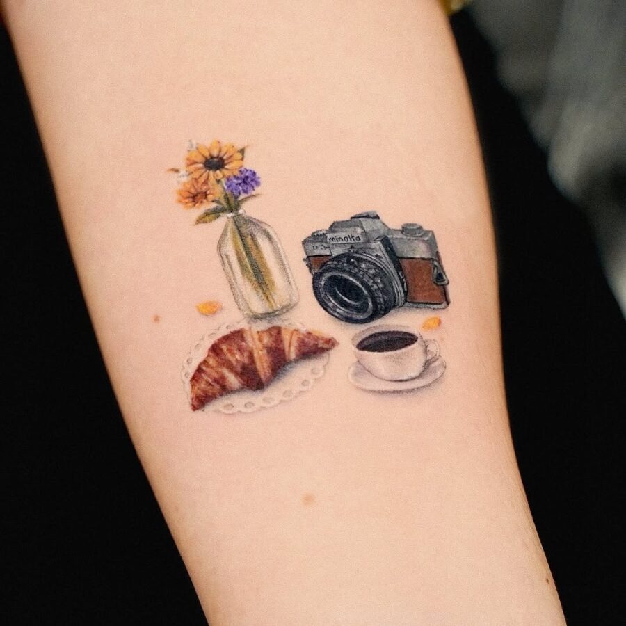 This tattoo captures a charming scene of a vintage camera alongside a delicious croissant, a cup of coffee, and flowers in a vase.