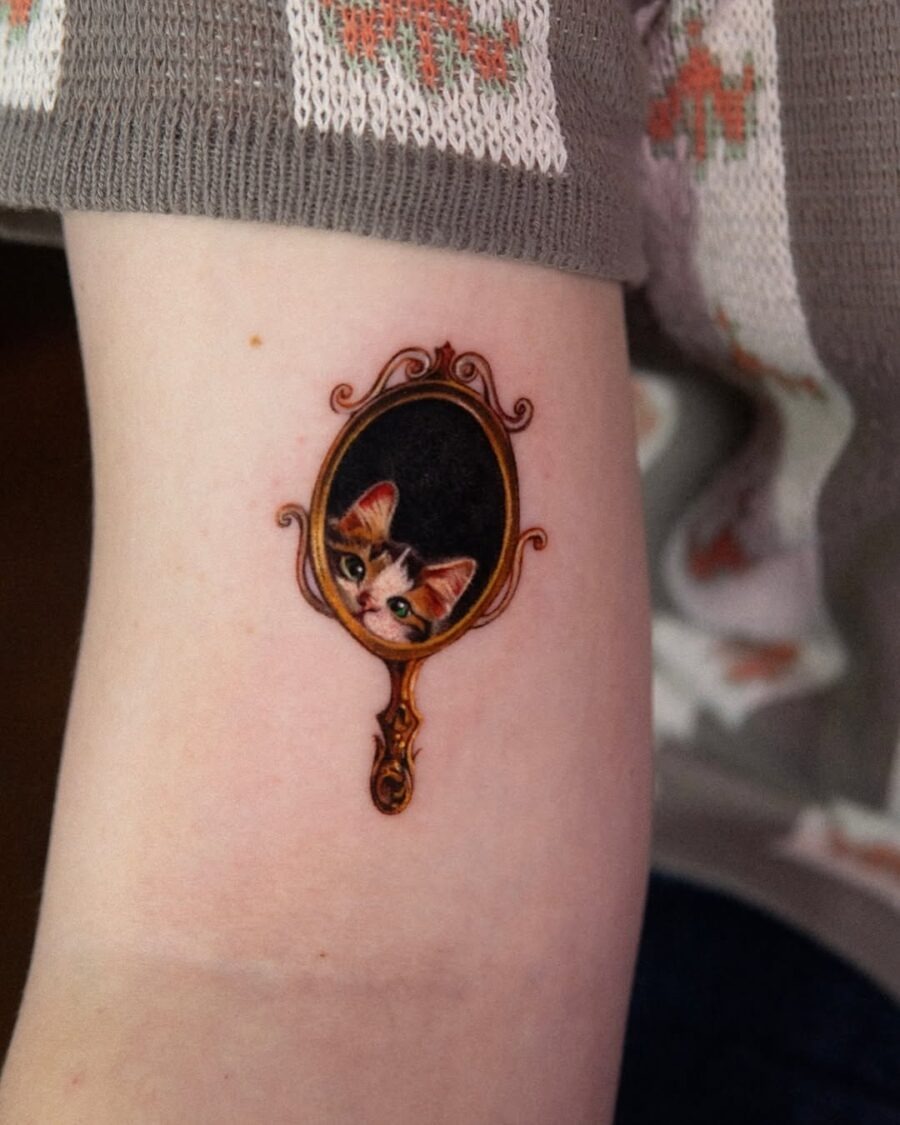 This tattoo features a charming cat peeking from a vintage mirror frame.