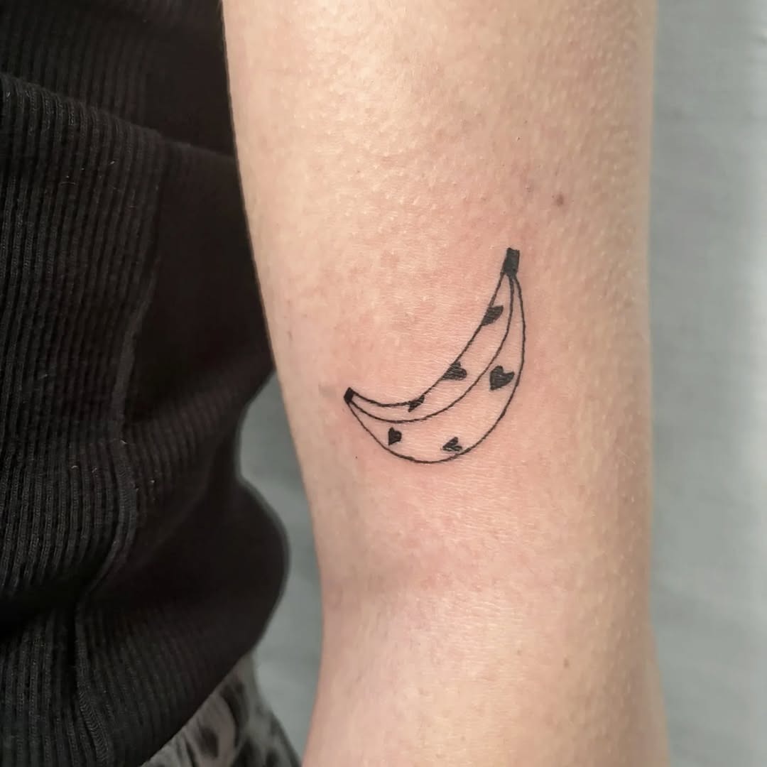 This cute banana tattoo features a simple outline adorned with tiny hearts, creating a playful yet sophisticated vibe.