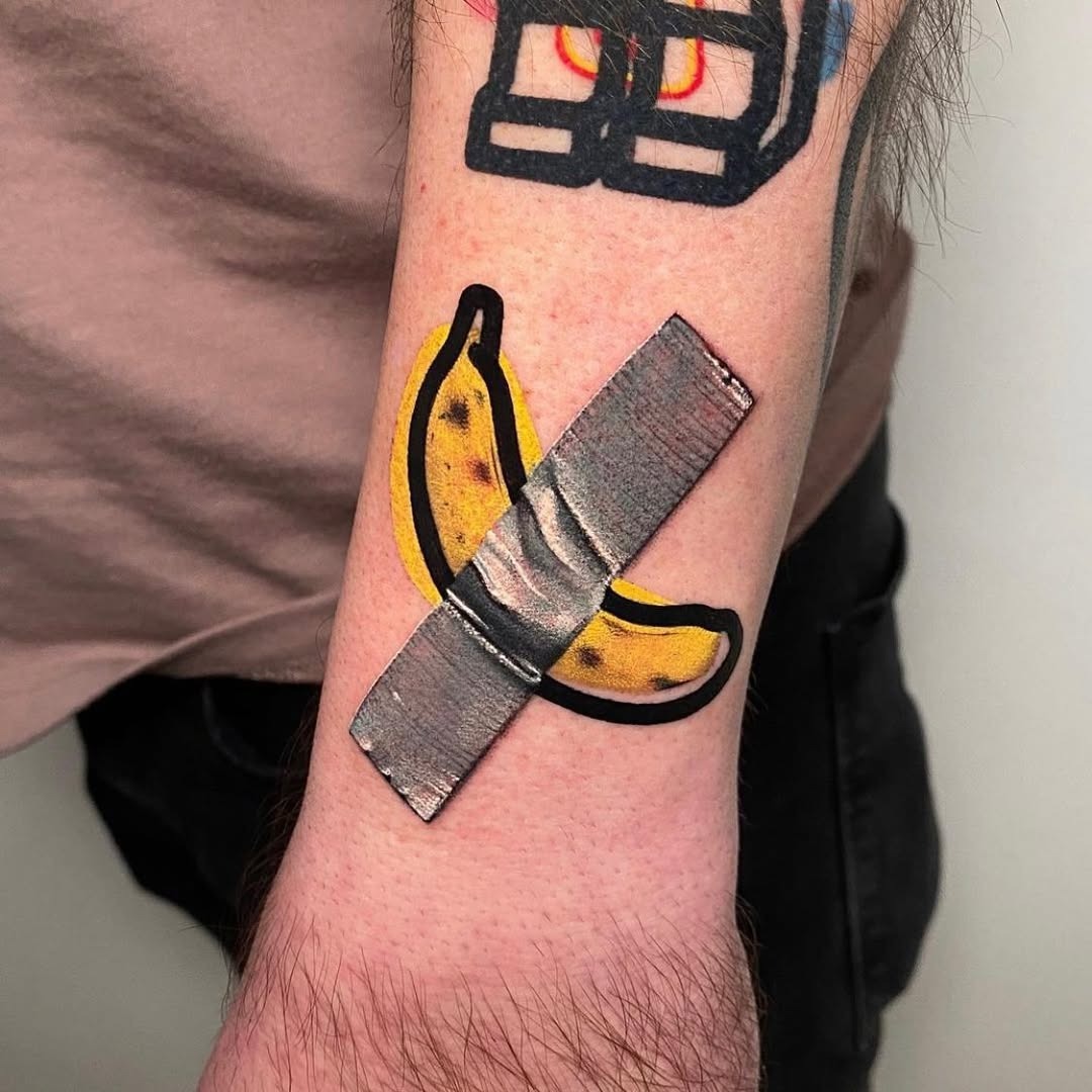 This playful banana tattoo features a metallic bandage over it, giving it a whimsical touch.