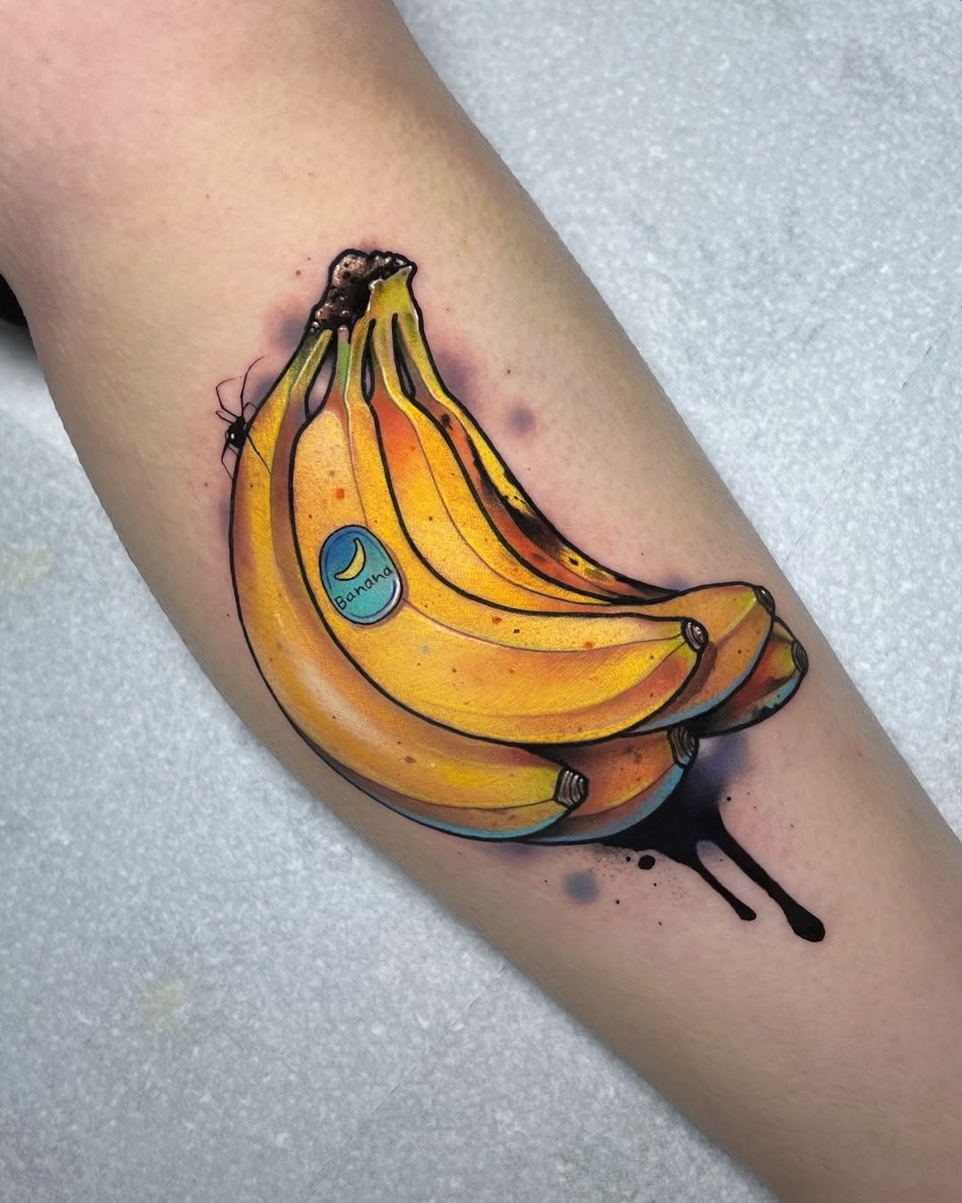 This striking tattoo showcases a bunch of bananas, complete with a little green sticker depicting the word 'banana.' The colors pop against the skin, giving it a playful and lively vibe.