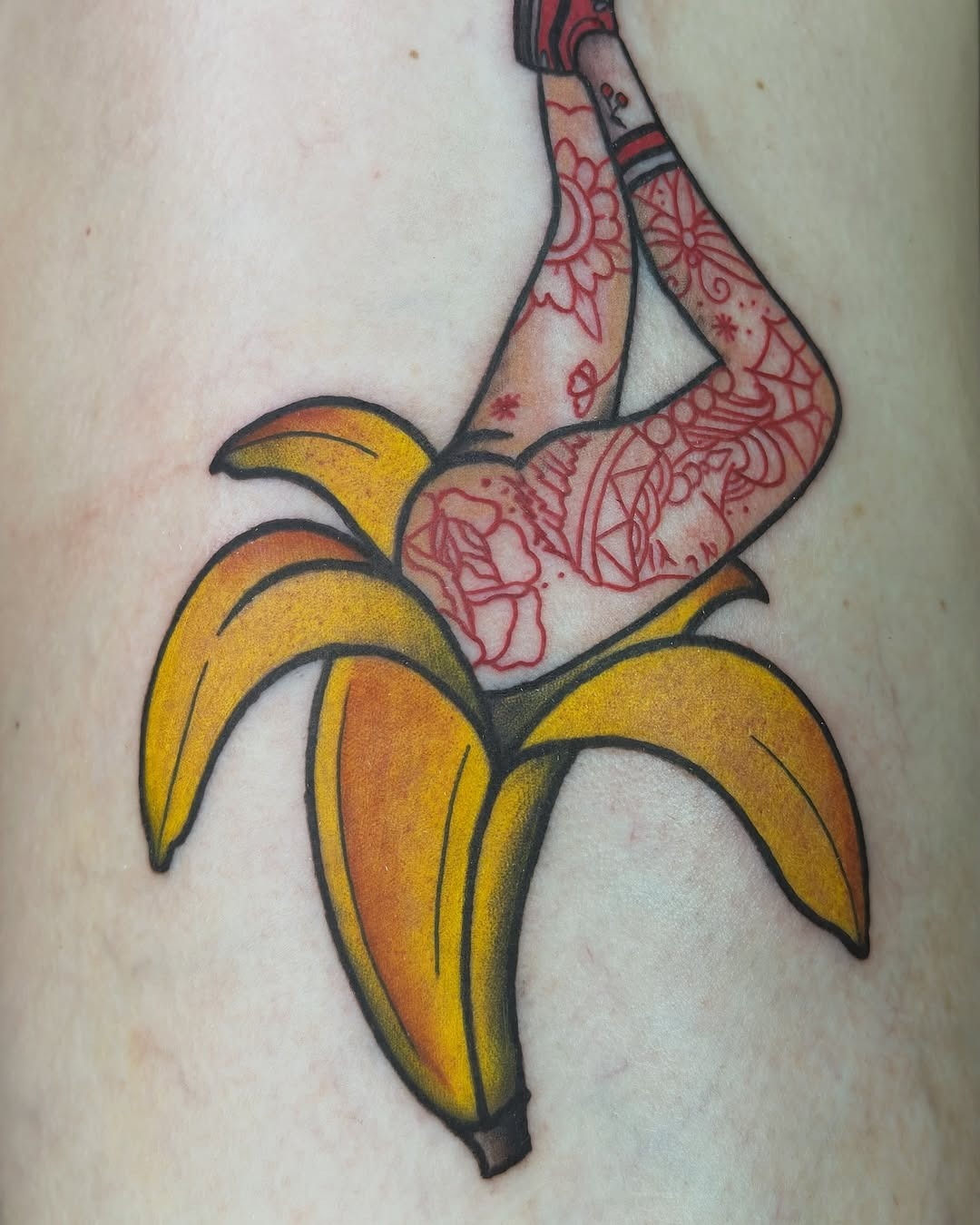 This tattoo features a playful design of a banana with a woman's legs emerging from it, adorned with intricate red floral patterns.