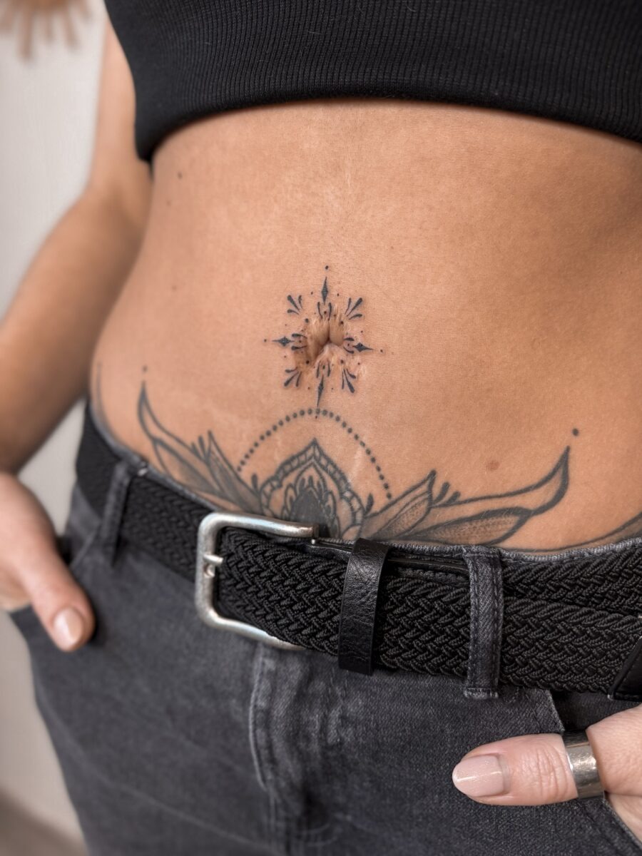 This belly tattoo features an elegant bloom at the center, radiating intricate lines and dots.