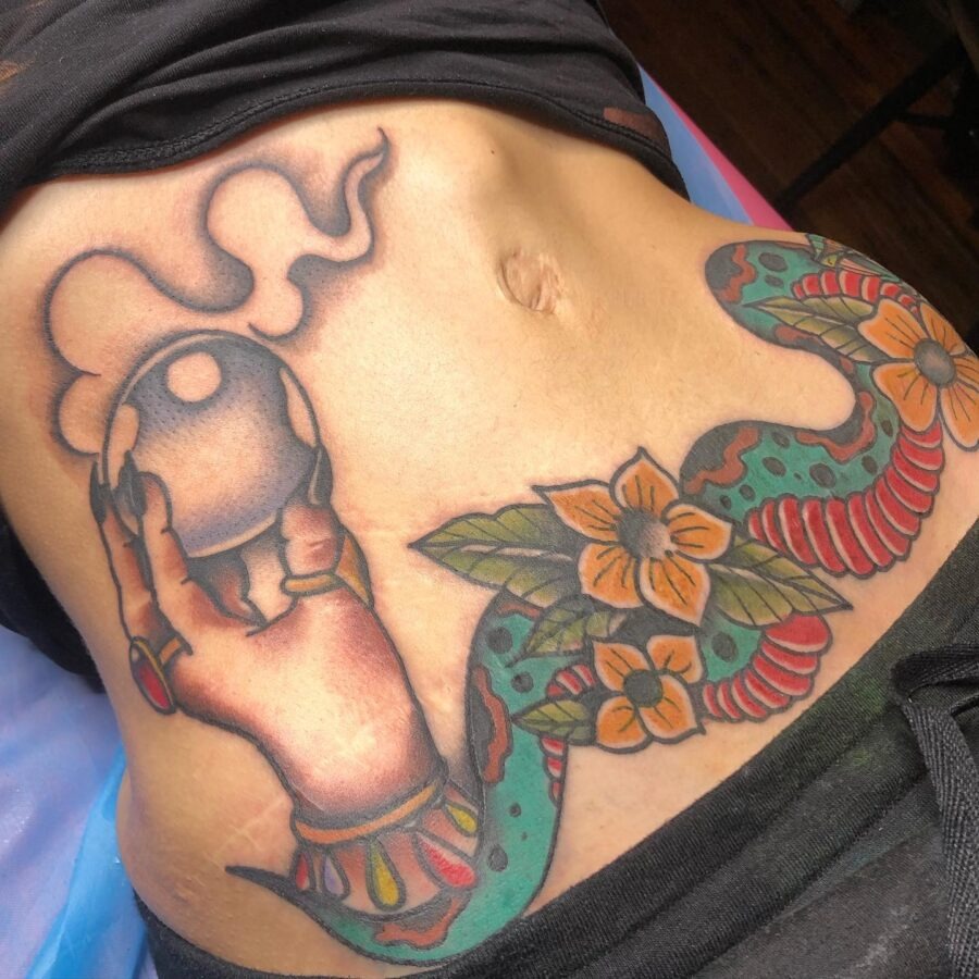 This striking belly tattoo features a hand holding a crystal orb, entwined with a snake and blooming flowers.