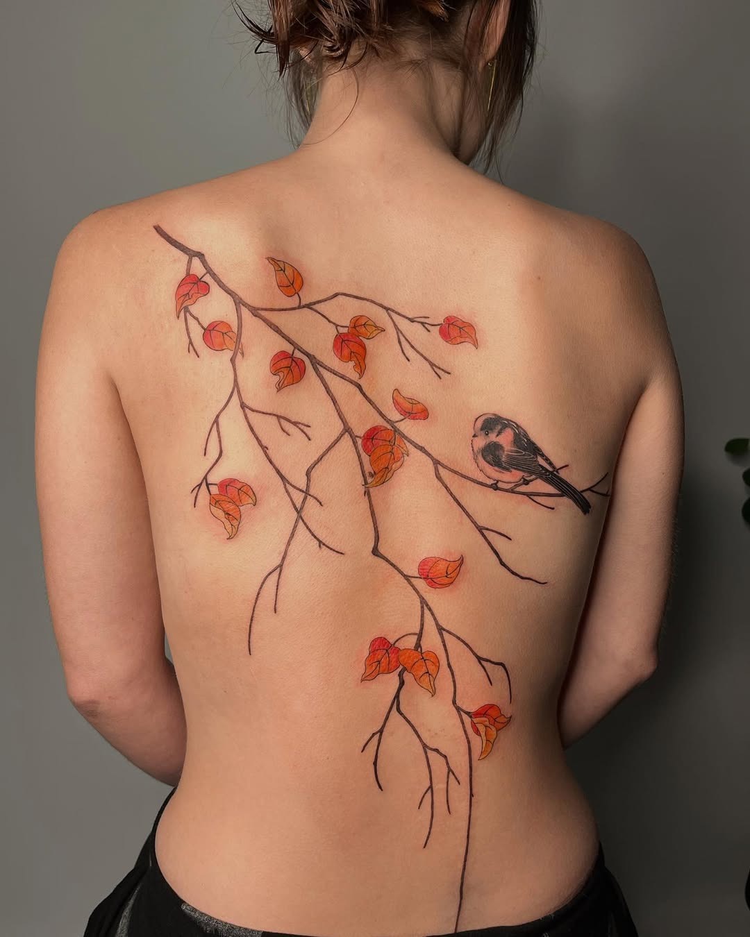 This captivating tattoo features a delicate birch tree branch adorned with red leaves and a charming bird.