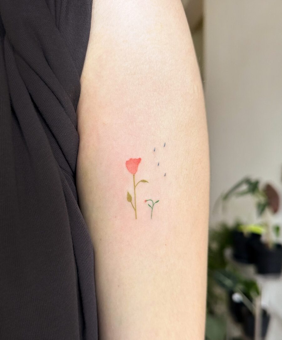 This tattoo showcases a delicate red flower with soft green leaves, set against a light background.
