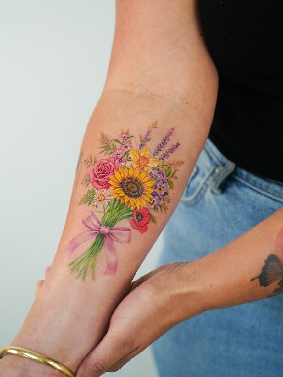 This stunning bouquet tattoo features a mix of flowers, including sunflowers, roses, and lavender, elegantly tied with a soft pink ribbon.