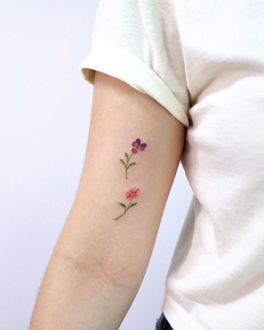 This charming tattoo showcases two beautiful flowers, a violet and a pink bloom, gracefully inked on the inside of the arm.