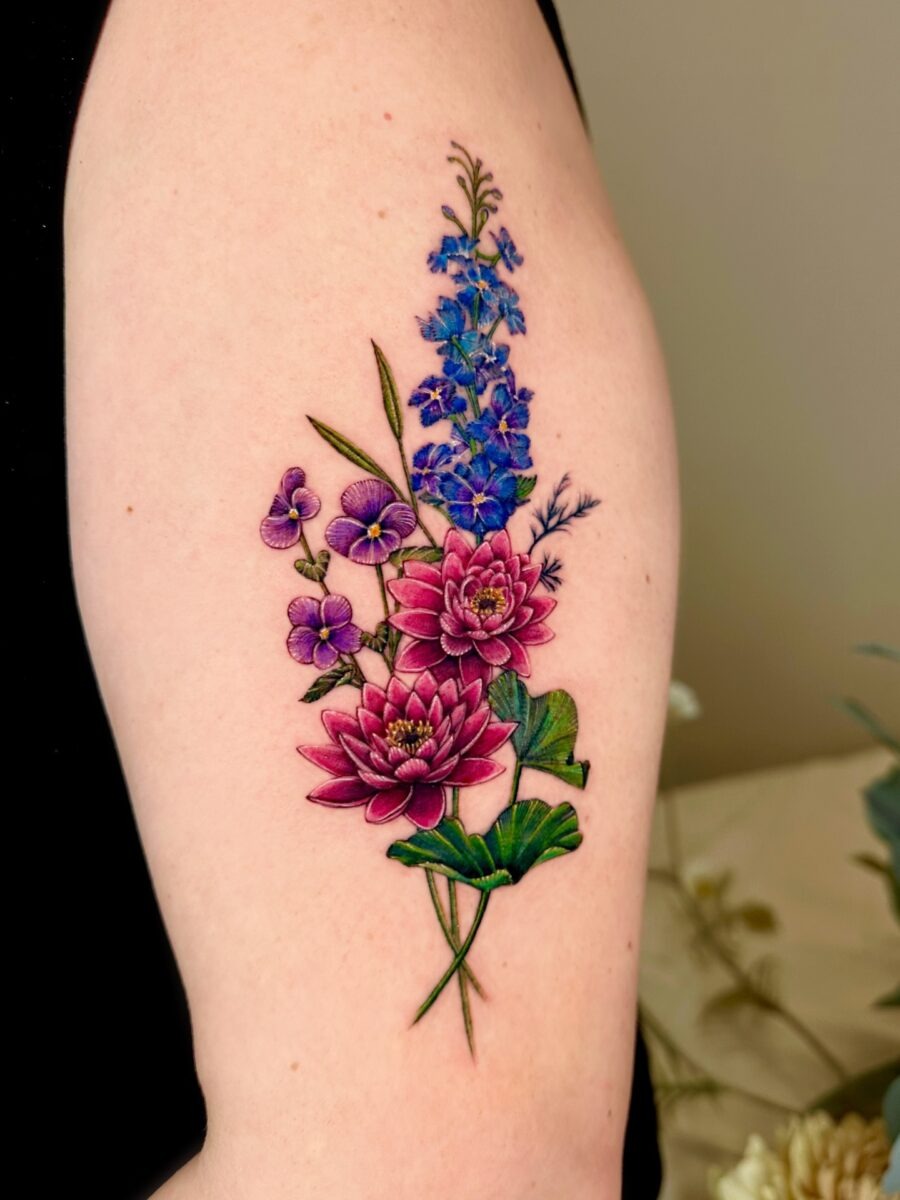 This beautiful tattoo features a lotus flower complemented by delicate blue delphiniums and purple violets.