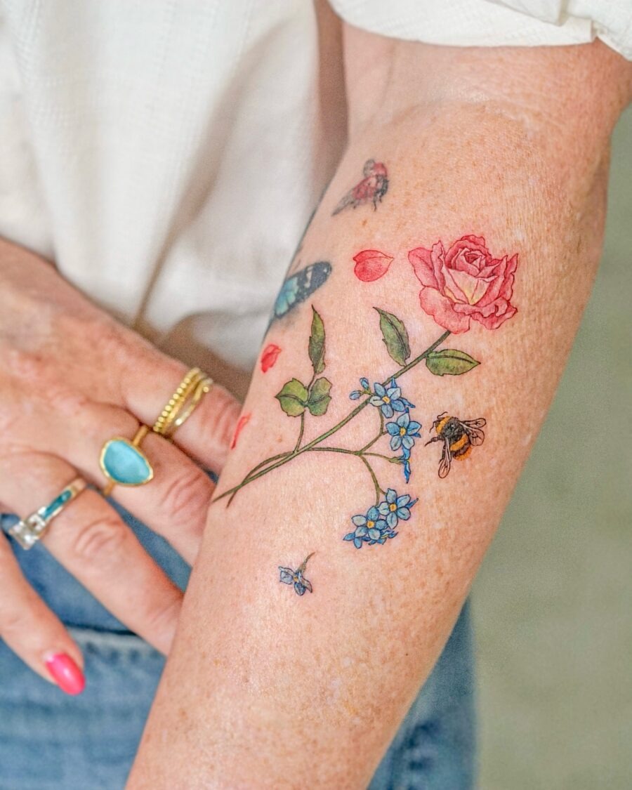 This tattoo beautifully showcases a rose, surrounded by delicate blue flowers and enchanting insects.