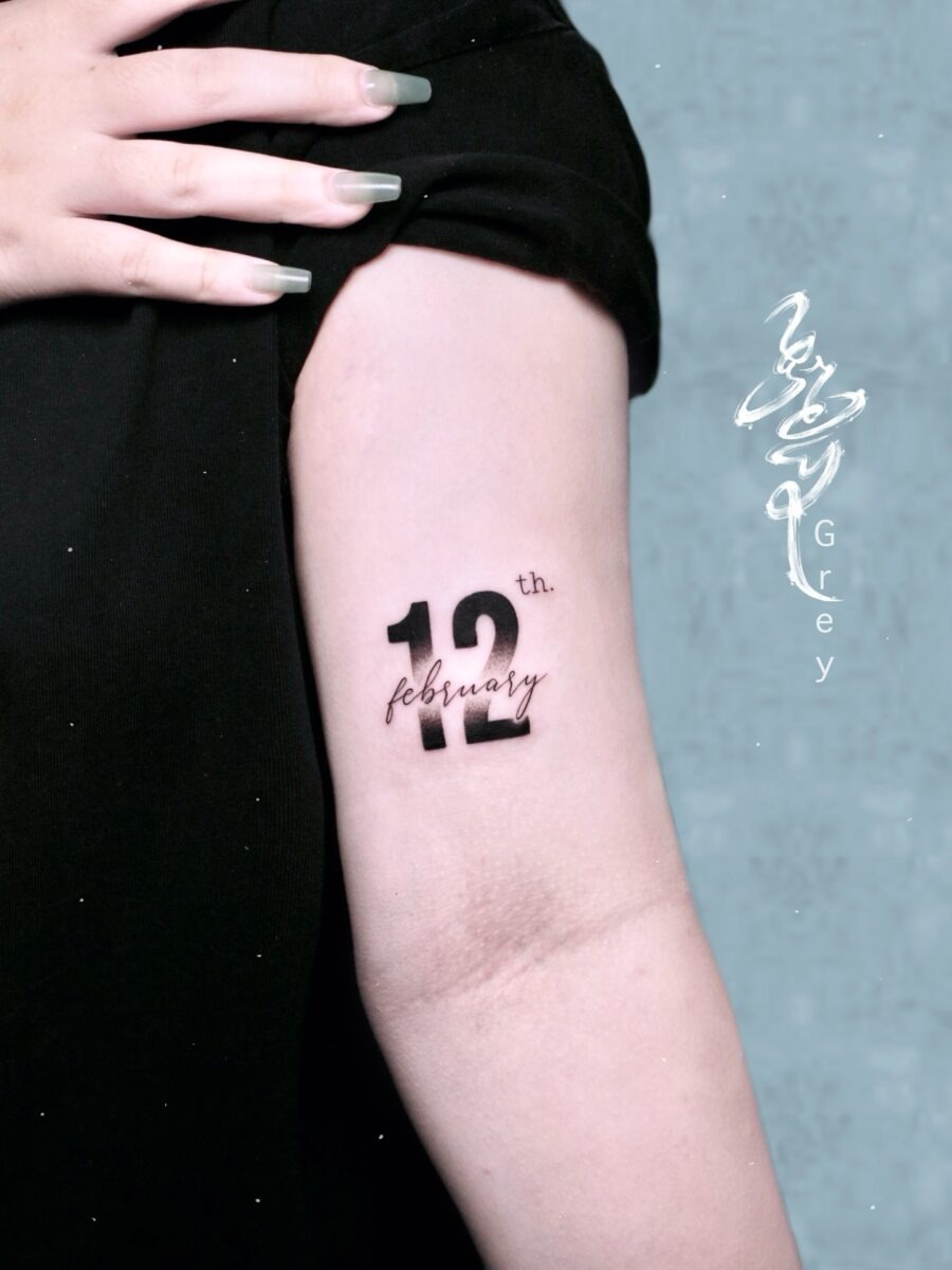 This birthday tattoo features a beautifully scripted date, '12th February,' inked on the upper arm.