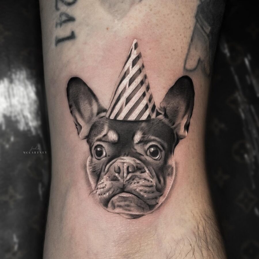 This tattoo features a realistic French Bulldog wearing a striped party hat.