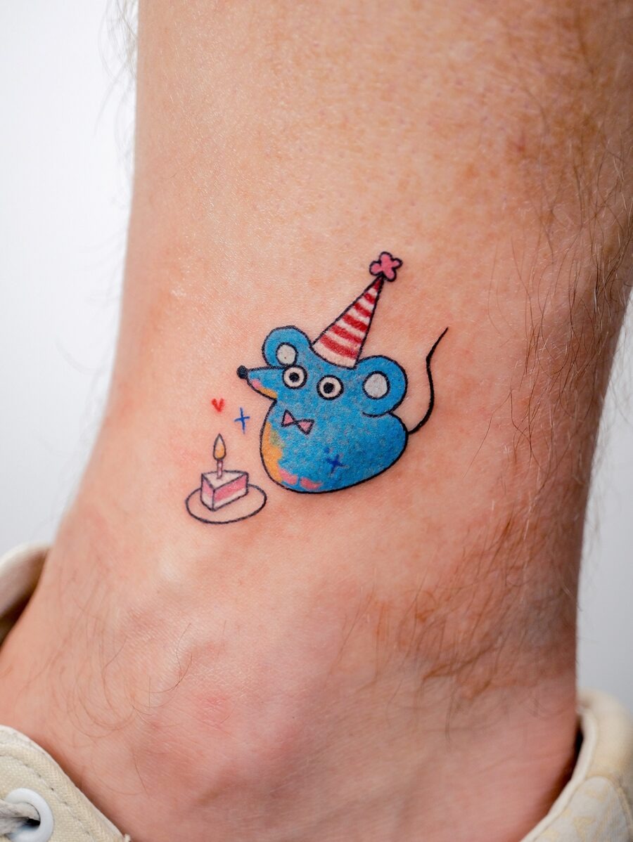 This adorable tattoo features a colorful mouse wearing a festive party hat, sitting beside a birthday cake.