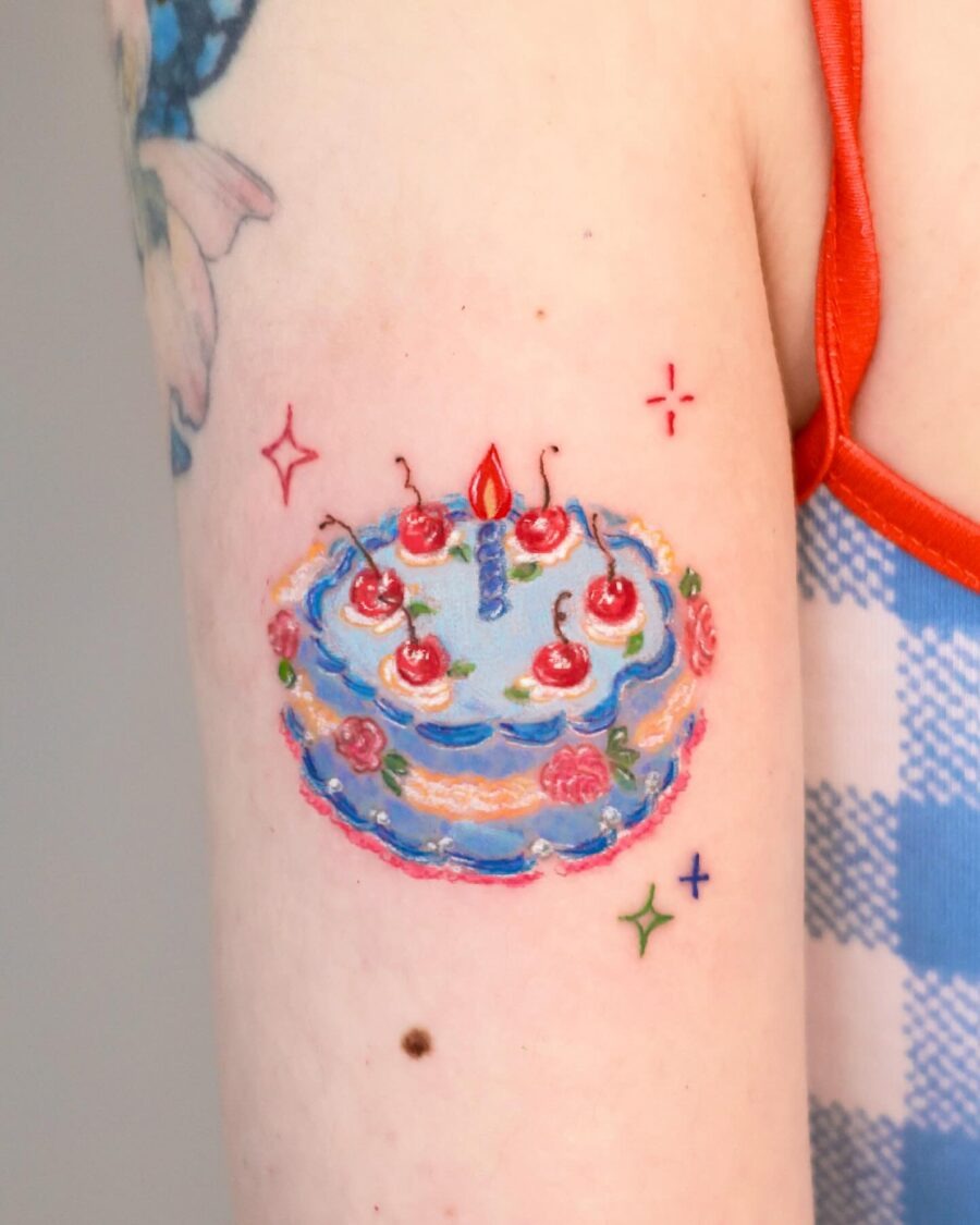 This tattoo features a colorful birthday cake adorned with cherries and flowers, radiating a festive vibe.