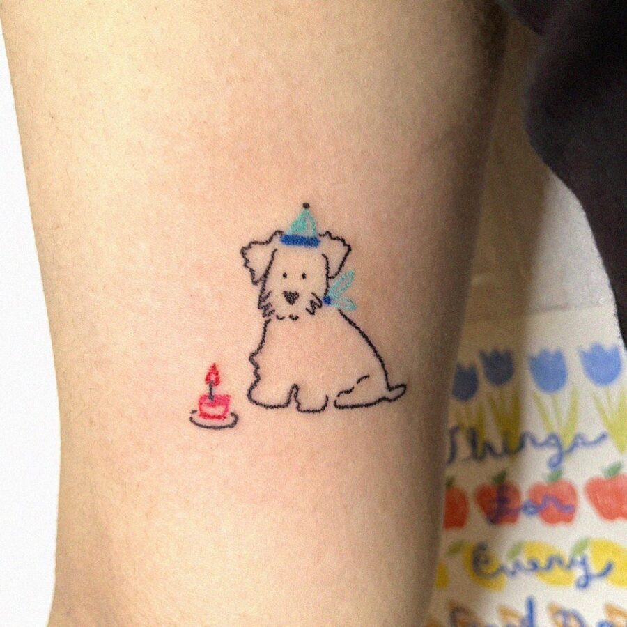 This tattoo features a charming dog wearing a party hat, sitting next to a small birthday cake.