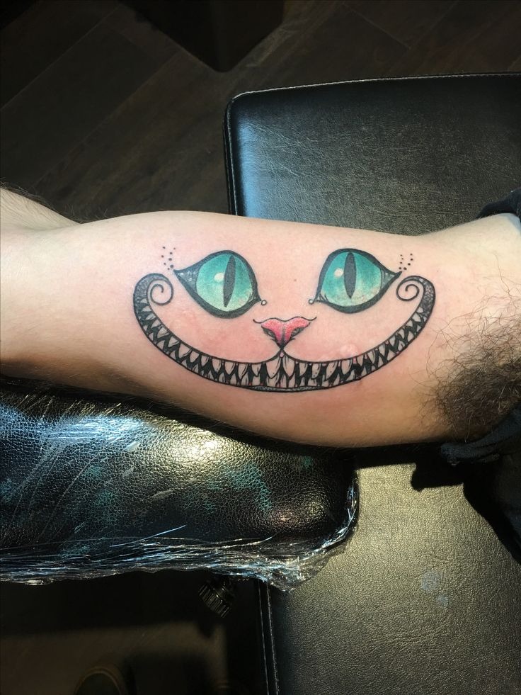 This tattoo focuses on the Cheshire Cat's wide grin and hypnotic eyes, rendered in a quirky, almost stencil-like style.