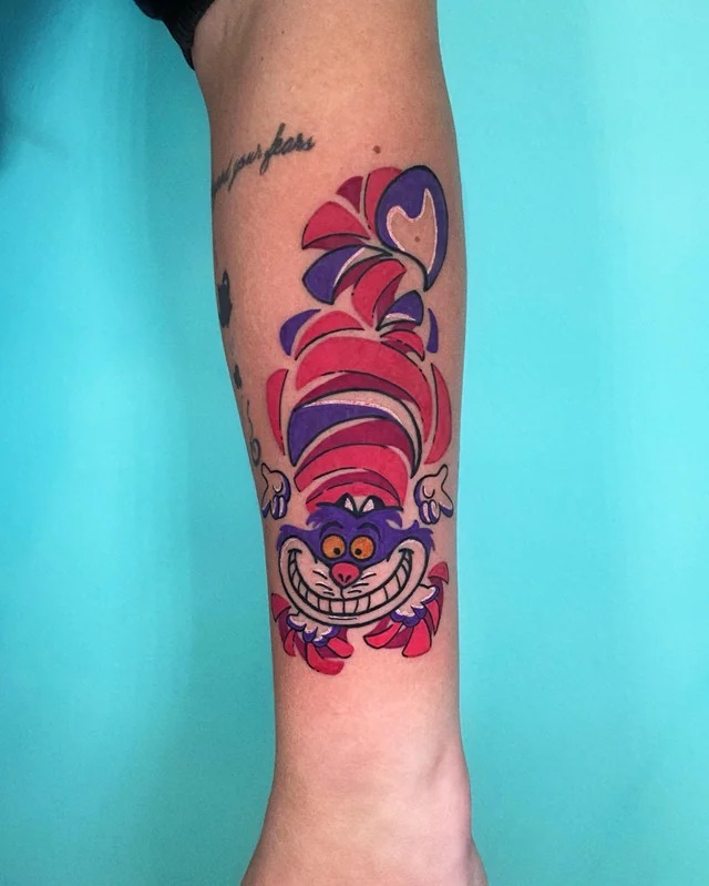 This tattoo features a, colorful depiction of the Cheshire Cat in a playful, cartoonish style.