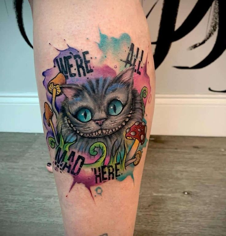 This watercolor tattoo is a explosion of colors surrounding the iconic Cheshire Cat and the famous phrase, "We're all mad here." The cat's fur is depicted in a soft gradient of blues and purples, blending seamlessly with the abstract background.