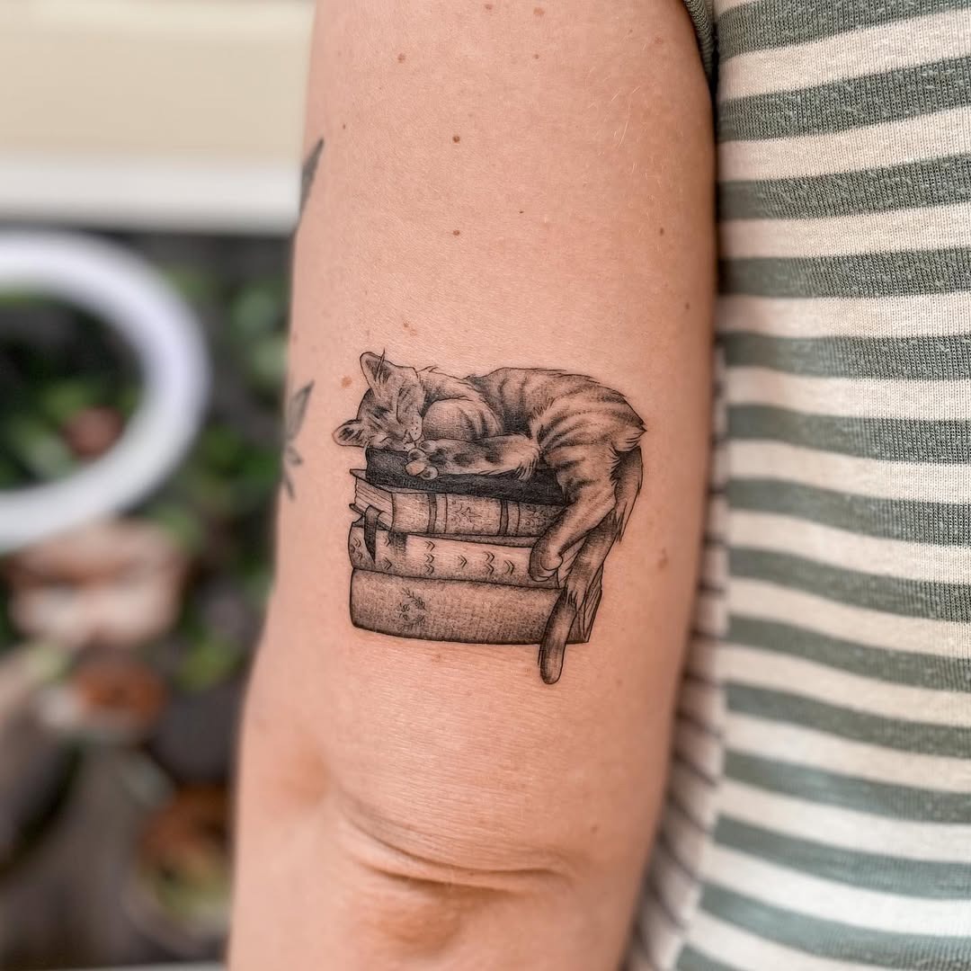 This tattoo showcases a delightful cat peacefully napping atop a stack of books.