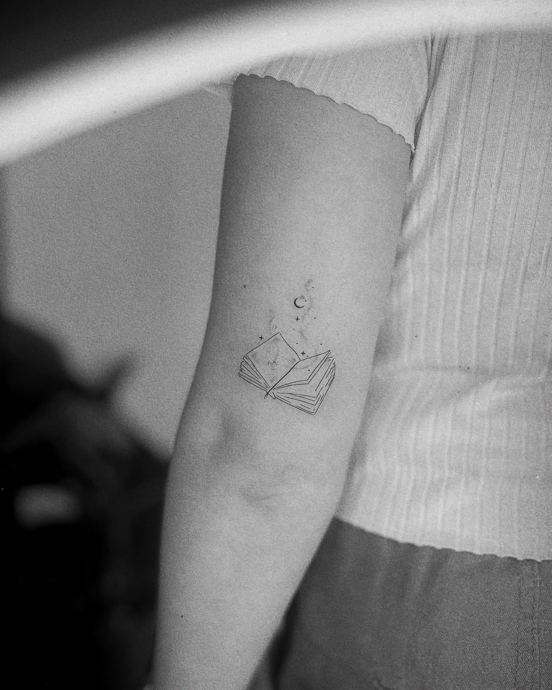 This tattoo features an open book surrounded by whimsical stars and a crescent moon.