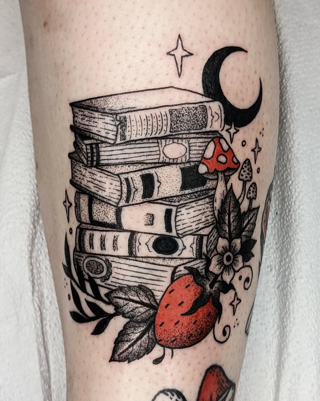 This tattoo showcases a charming stack of books adorned with flowers, mushrooms, and a crescent moon, creating a whimsical vibe.
