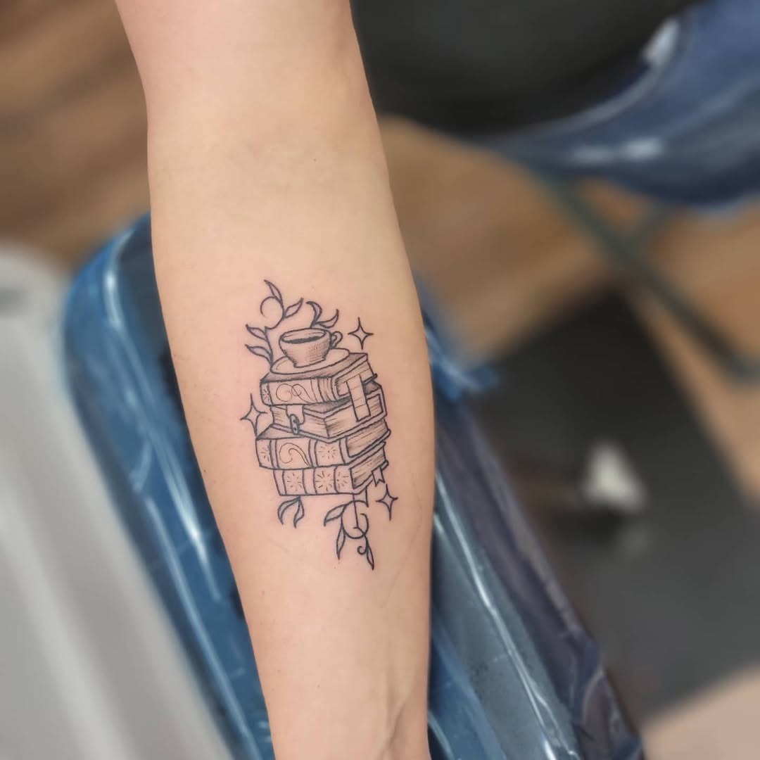 This tattoo features a whimsical stack of intricately detailed books topped with a delicate teacup, surrounded by floral elements.