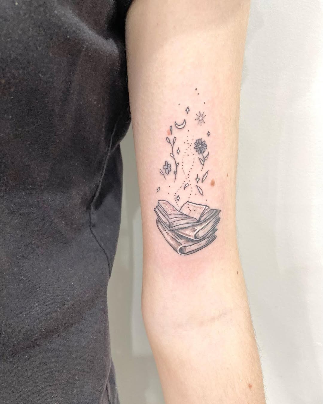 This tattoo features an intricately detailed open book, embellished with whimsical flowers and floating symbols.