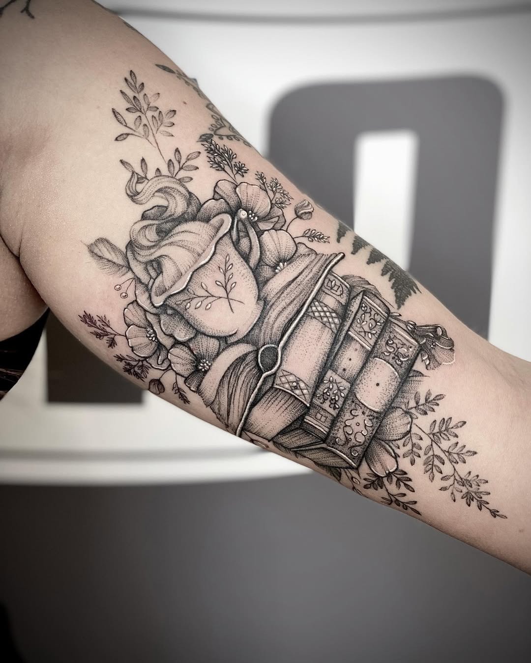 This tattoo features a beautifully detailed stack of books surrounded by delicate flowers.
