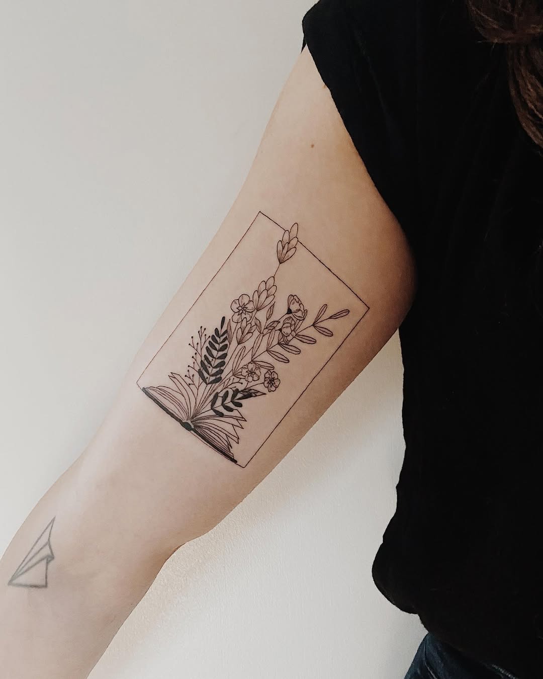 This tattoo features a beautifully detailed illustration of an open book adorned with delicate flowers and leafy elements.