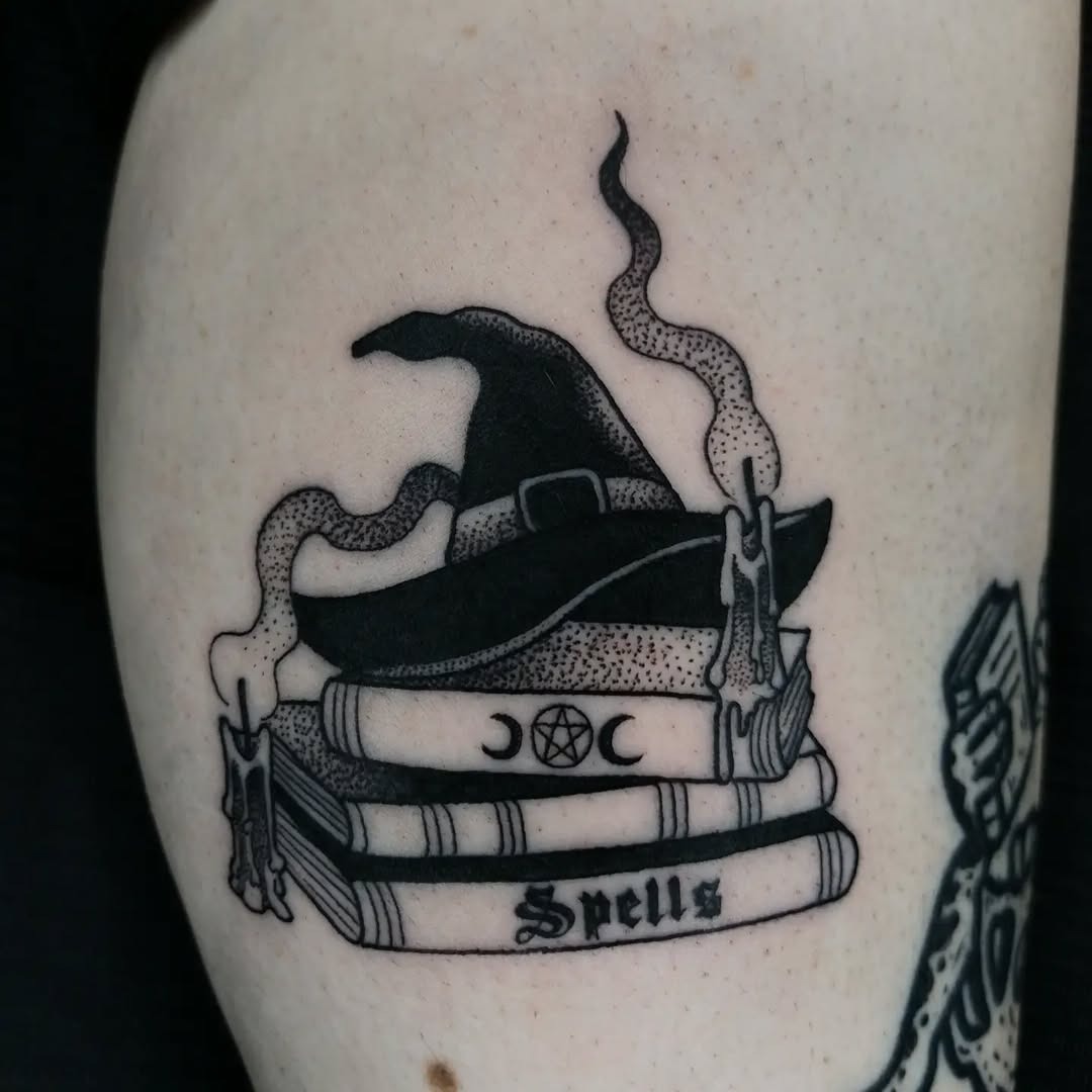 This tattoo features a whimsical witch's hat resting atop a stack of books, with the word 'Spells' elegantly inscribed.