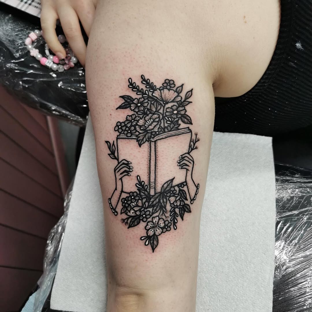 This tattoo features a pair of hands gracefully holding an open book, surrounded by beautiful floral designs.