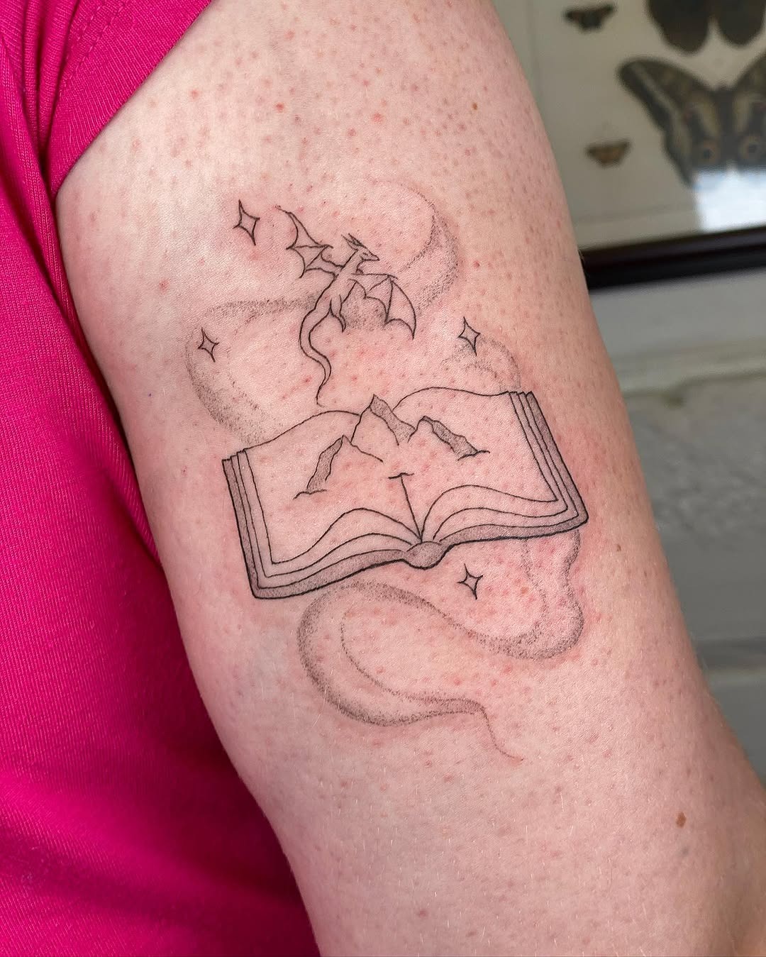 This enchanting tattoo features an open book with mountains, surrounded by a whimsical dragon.