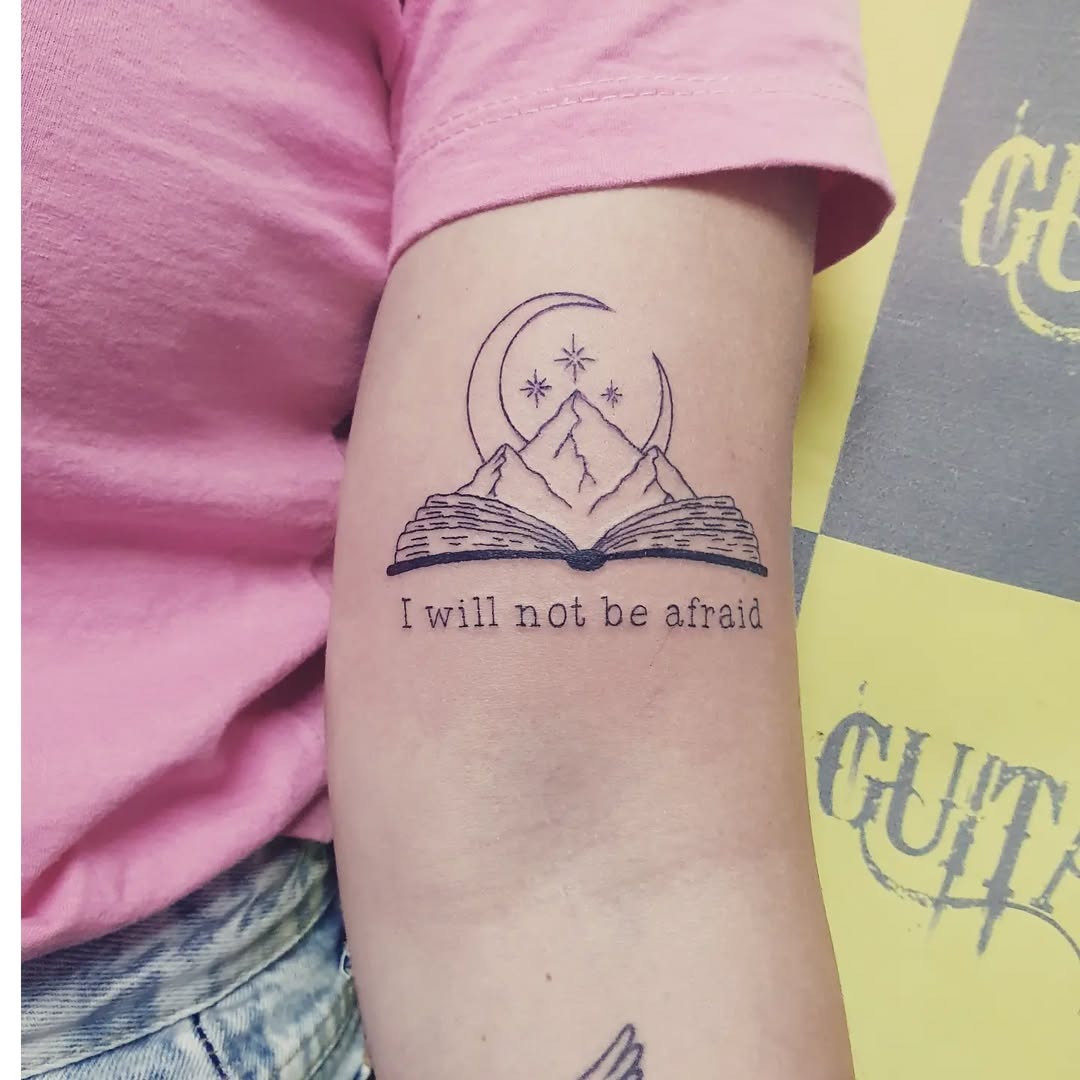 This tattoo features an open book with a serene mountain landscape and a celestial design above.