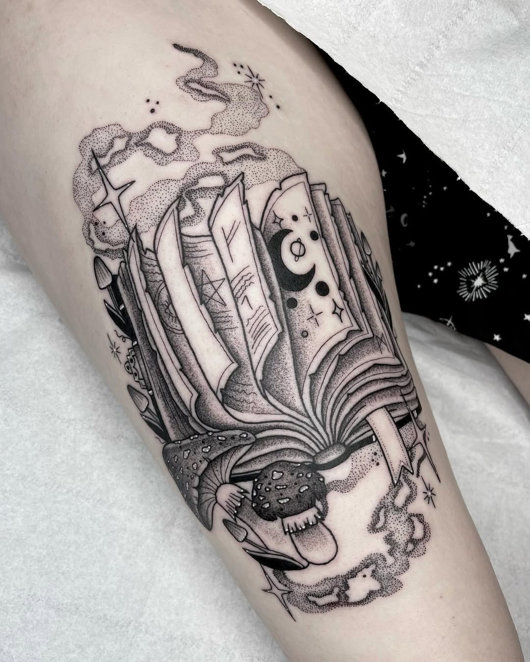 This tattoo features a beautifully detailed open book surrounded by celestial elements like stars and clouds.