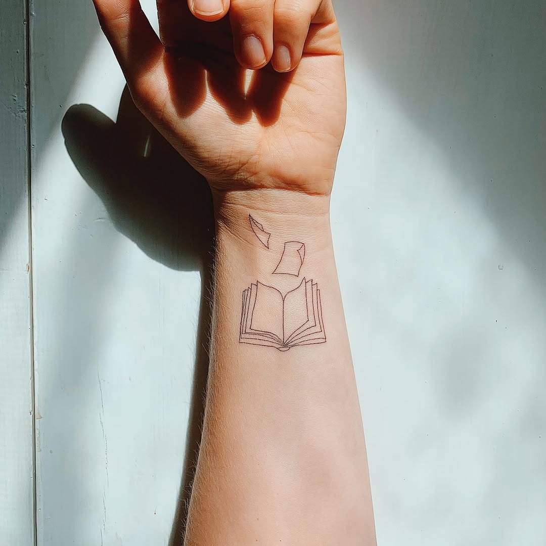 This tattoo features a minimalist depiction of an open book with a few pages fluttering above it.