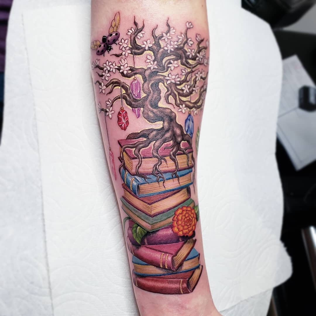 This tattoo showcases a tree with intricate roots intertwined with colorful books.