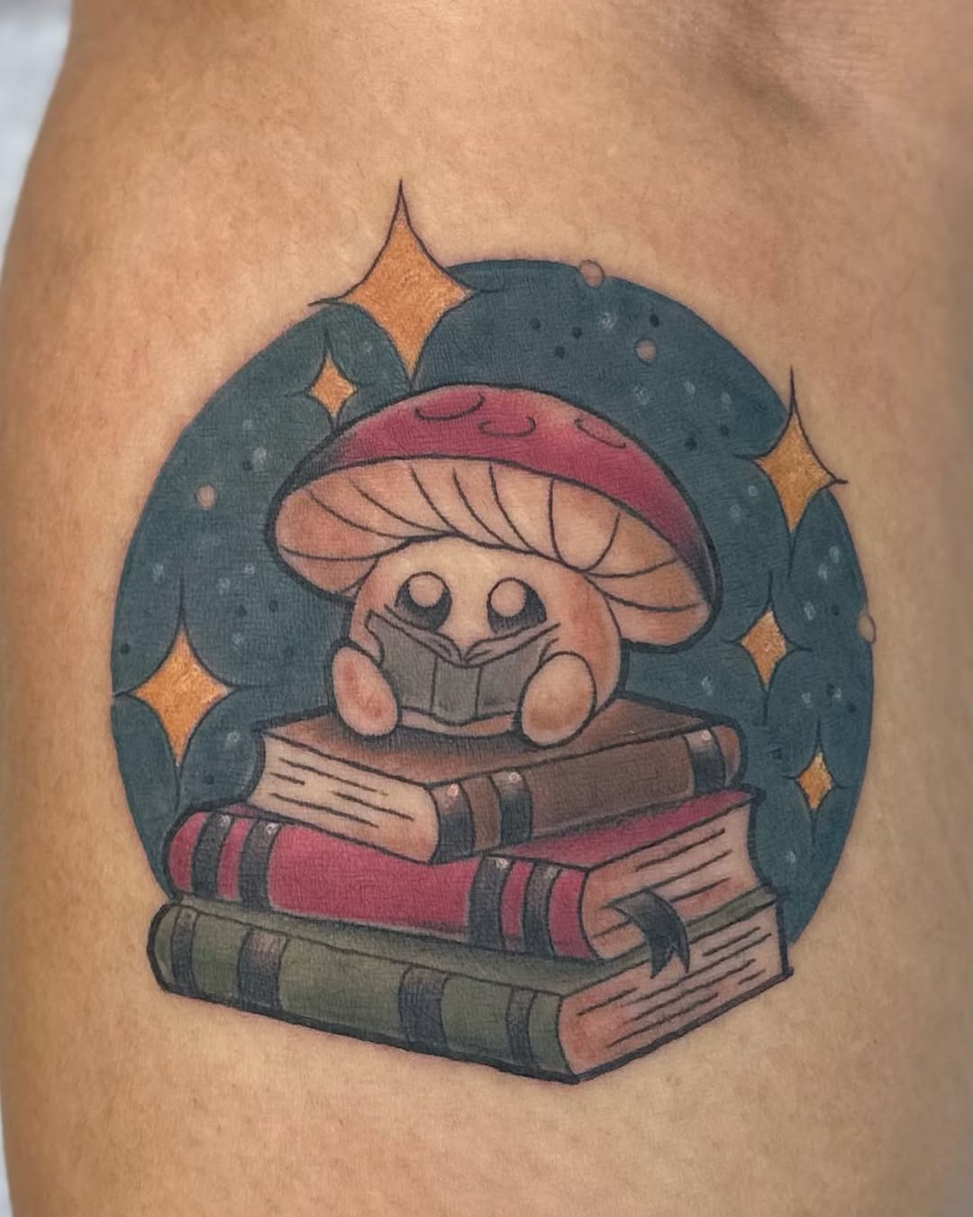 This charming tattoo features an adorable mushroom character engrossed in a book, sitting atop colorful stacked volumes.