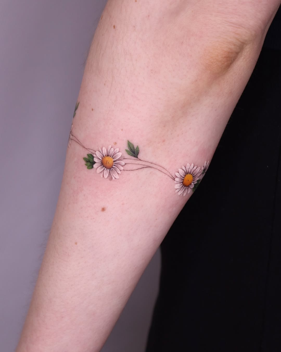 This charming bracelet tattoo features a delicate line of daisies intertwined with green leaves, wrapping gracefully around the forearm.