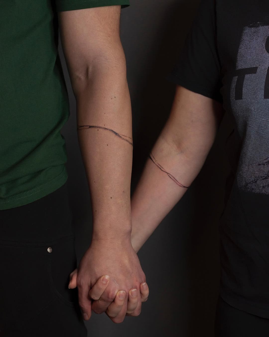 This image features two individuals holding hands, each adorned with a minimalist bracelet tattoo around their wrist.