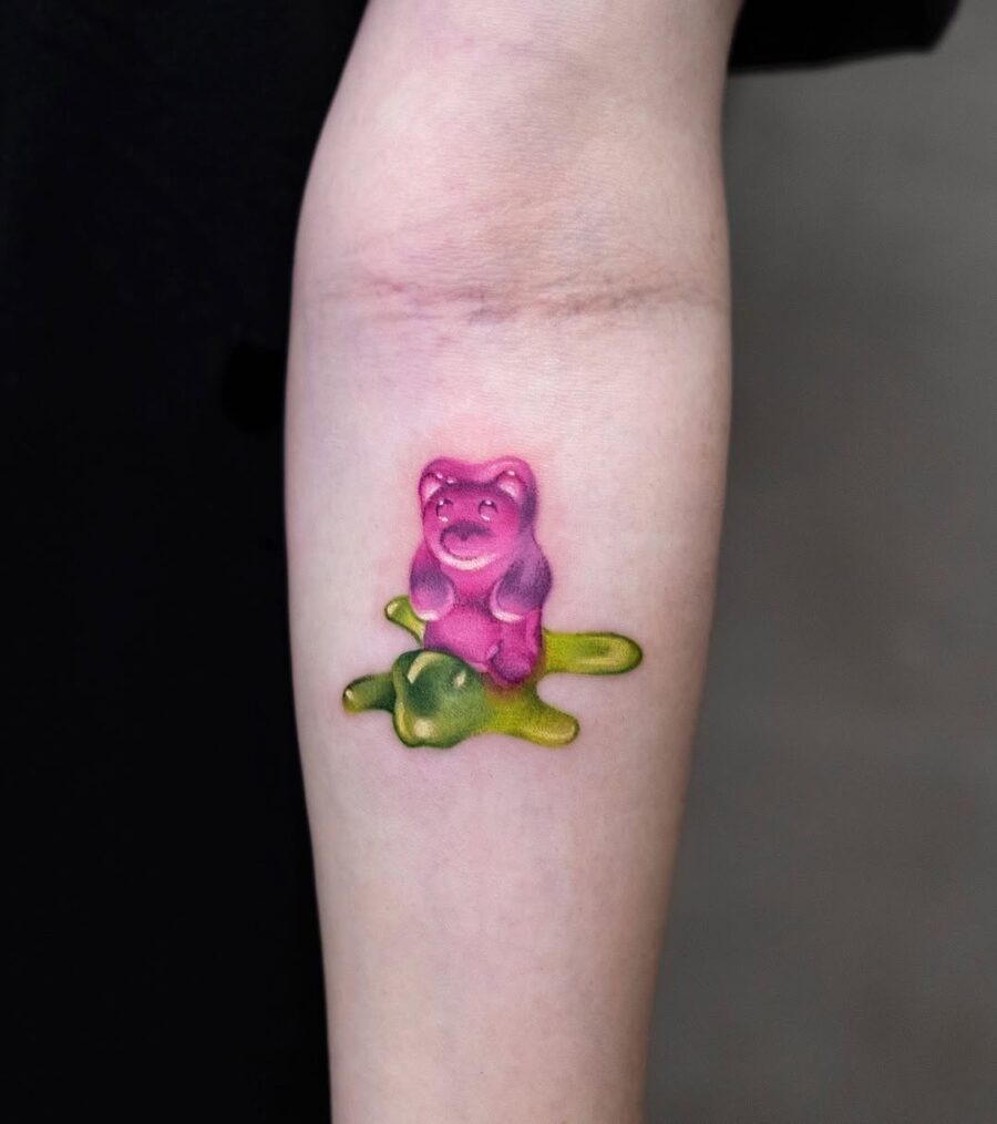 This playful tattoo features a vivid pink gummy bear sitting atop a green blob.