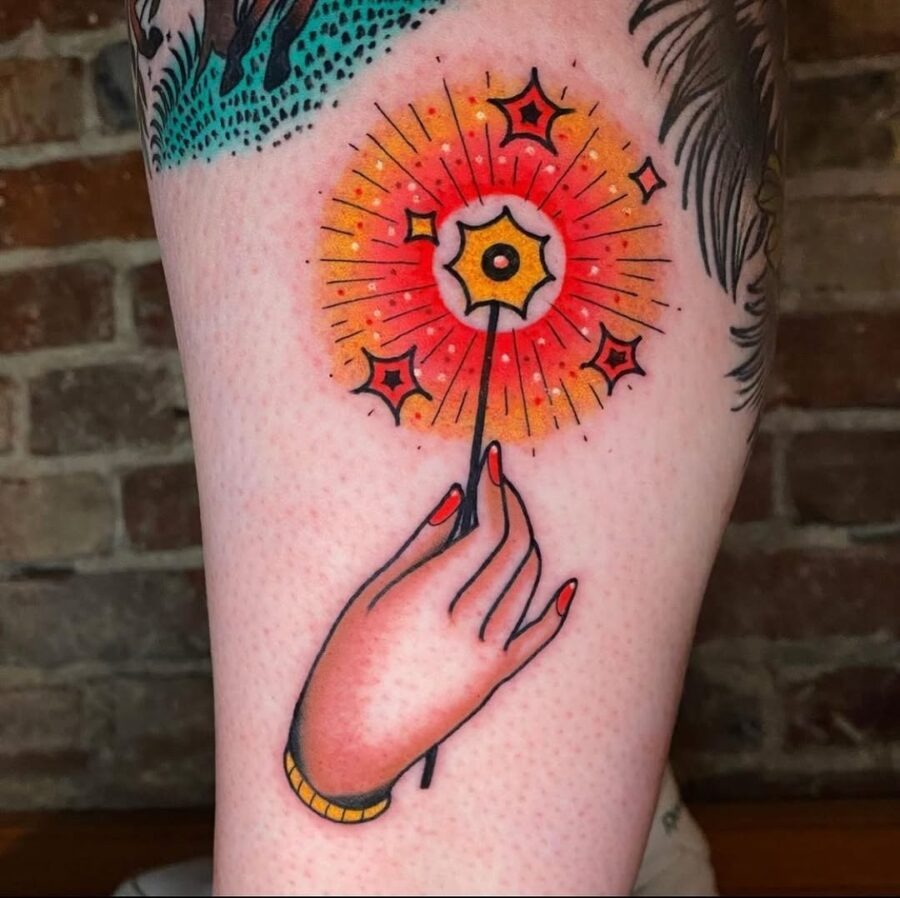 This tattoo features a beautifully detailed hand clutching a sparkler that radiates bright shades of orange and yellow.