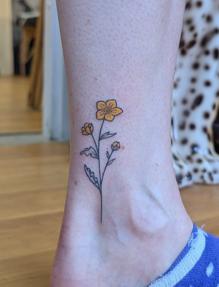 This ankle tattoo showcases a delicate buttercup flower design with yellow petals and detailed leaves.