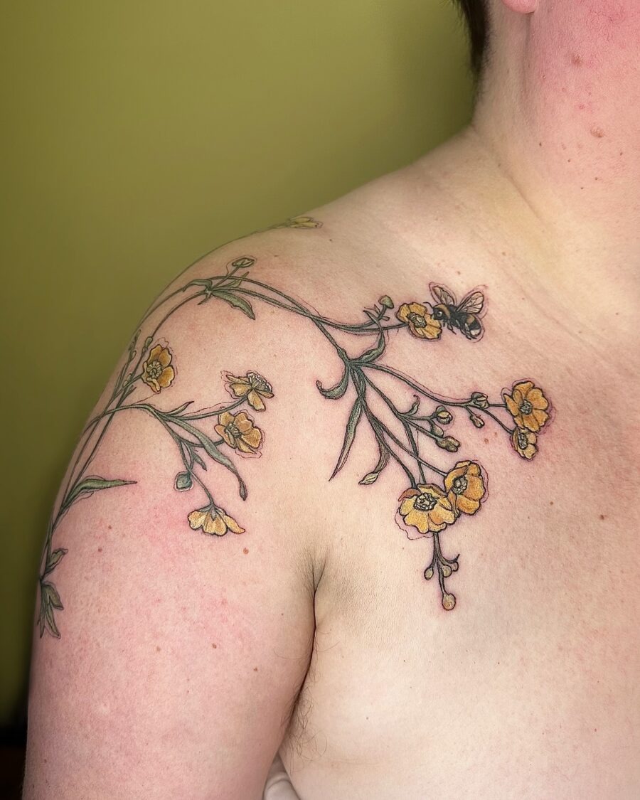 This tattoo features charming buttercup flowers gracefully cascading over the shoulder.