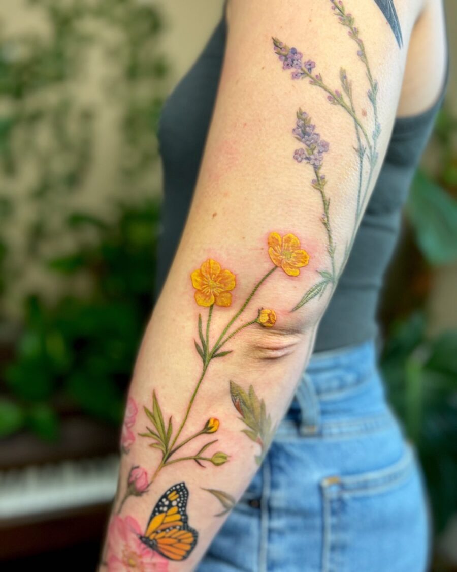 This beautiful arm tattoo features delicate buttercup flowers intertwined with colorful blooms and a butterfly.