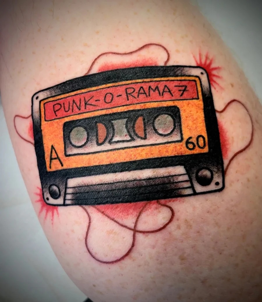 This tattoo features a classic cassette tape labeled 'PUNK-O-RAMA 7', highlighted with a splash of red ink.