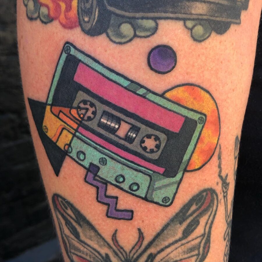 This tattoo showcases a cassette tape emerging from a box, reminiscent of nostalgic 80s aesthetics.