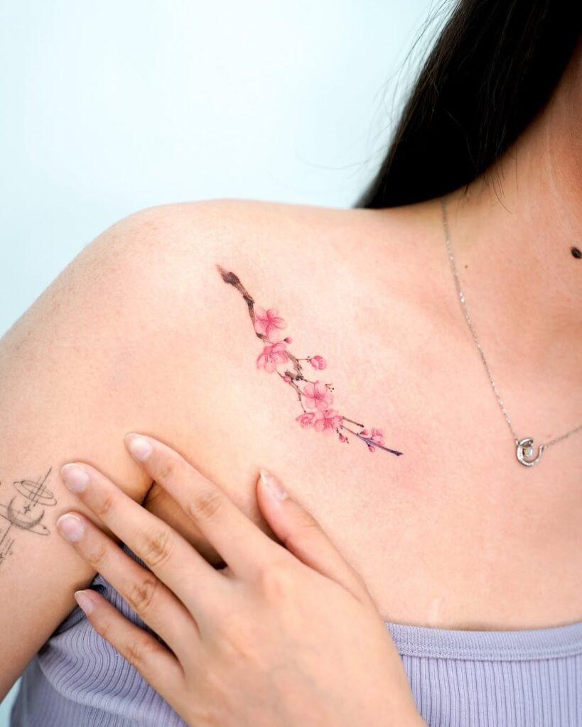 A graceful collarbone tattoo featuring soft pink cherry blossoms on a slender branch.