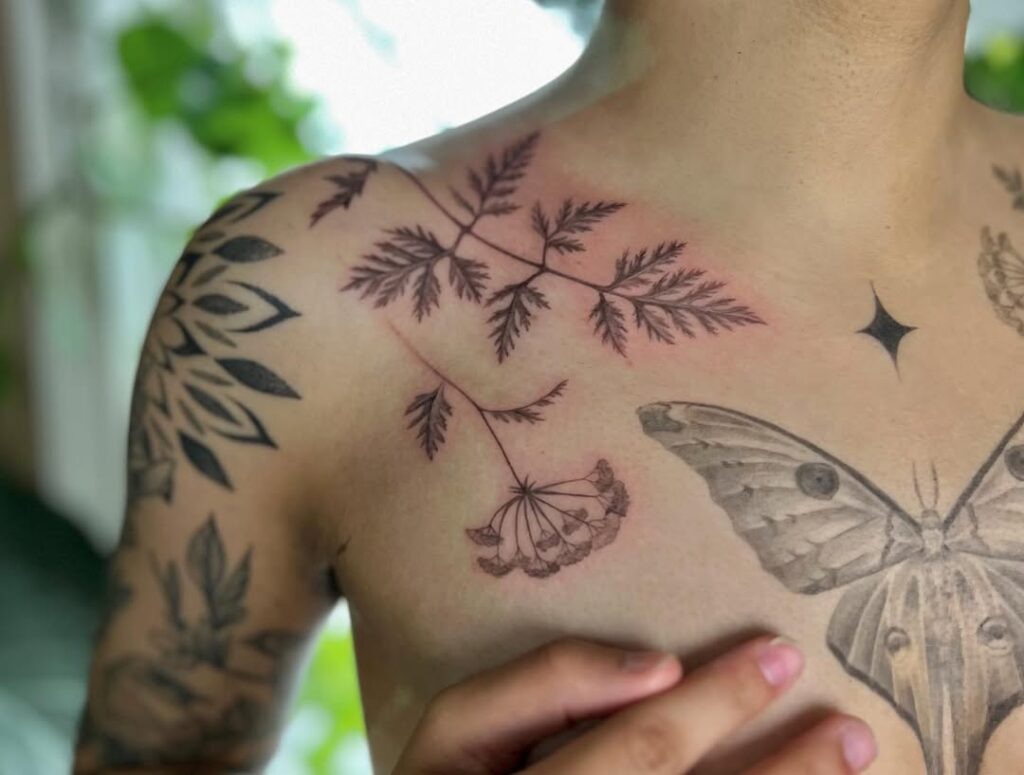 An elegant tattoo featuring detailed botanical leaves and stems extending across the collarbone.